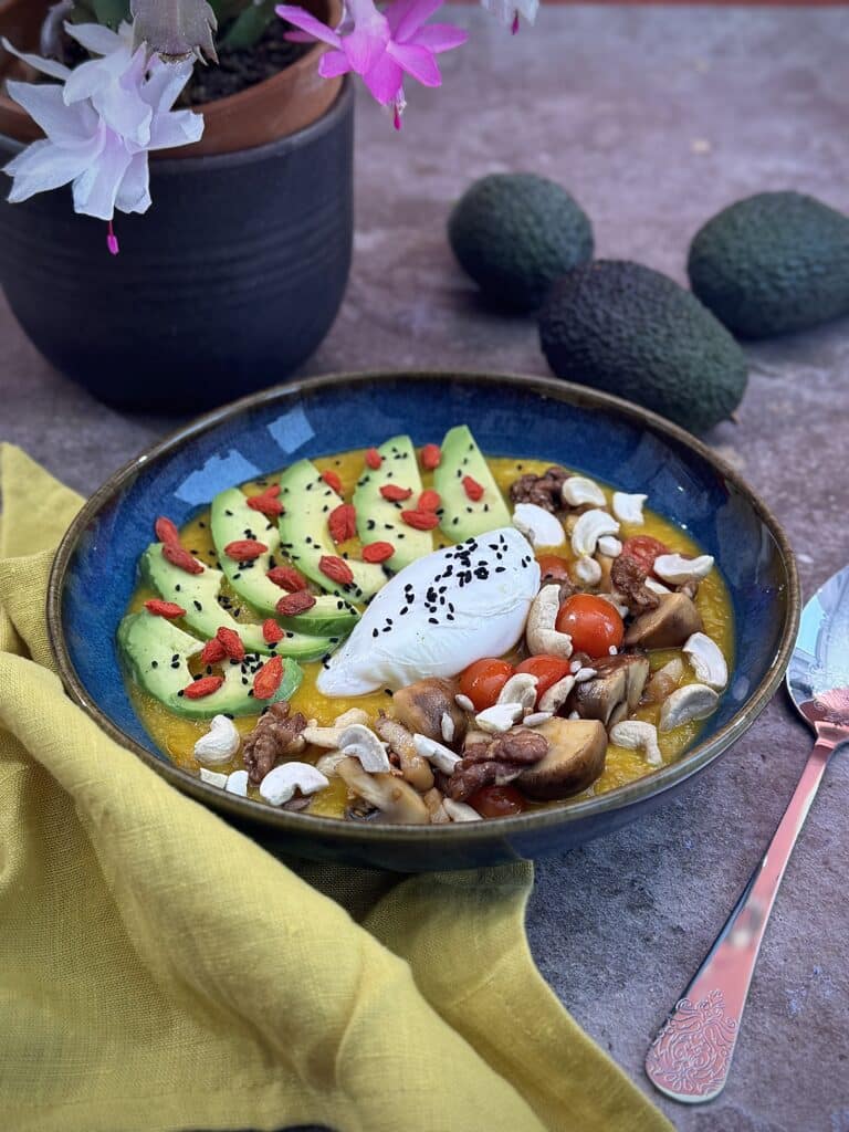 Pumpkin, potato, parsnip veloute in a dark blue bowl with poached egg on top, decorated with sliced avocado, goji berries, black cumin, nuts and seeds