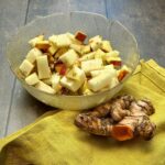 Side view of a bowl of kohlrabi apple salad with fresh turmeric slices and a turmeric root placed next to the salad.