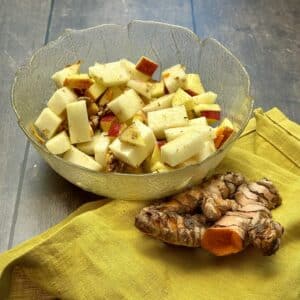 Side view of a bowl of kohlrabi apple salad with fresh turmeric slices and a turmeric root placed next to the salad.