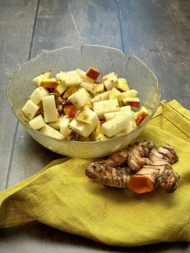Side view of a bowl of kohlrabi apple salad with fresh turmeric slices and a turmeric root placed next to the salad.