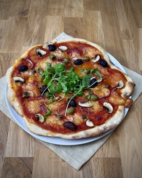 Overhead view of homemade wheat sourdough pizza topped with mushrooms, olives, capers, mozzarella and fresh rocket.