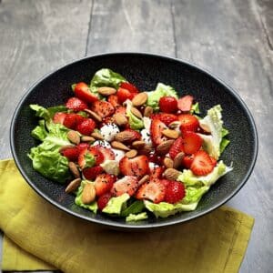 Side view of romana salad with strawberries, redcurrants, mozzarella and almonds in a black bowl.