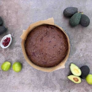 freshly baked avocado brownie cake in round baking form with avocados limes and raspberries around it