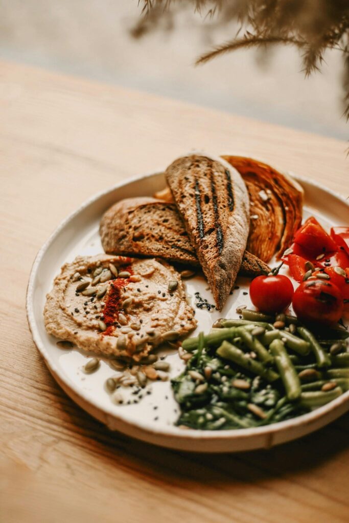 plate with hummus, toasted sourdough bread, green beans, leafy greens, seeds, and tomatoes as a simple fiber-friendly meal